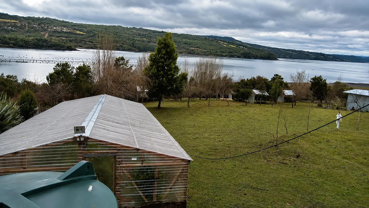Paisaje rural con invernadero, pradera y un gran cuerpo de agua rodeado de colinas